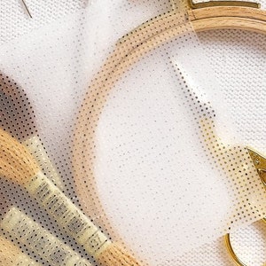 May include: A white fabric with a clear plastic sheet with a grid pattern on top. The plastic sheet is being used to help with embroidery. There are embroidery floss spools and a wooden embroidery hoop in the image.