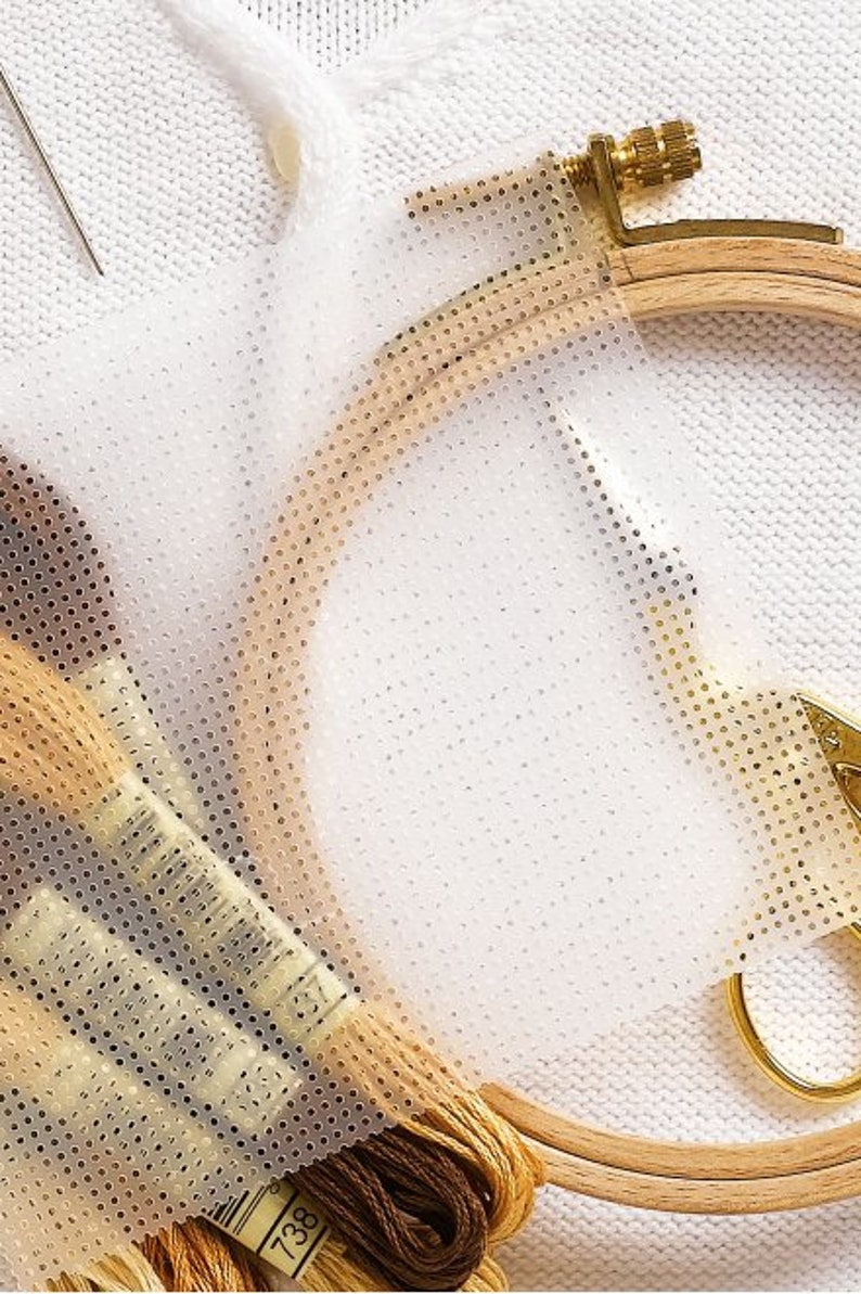 May include: A white fabric with a clear plastic sheet with a grid pattern on top. The plastic sheet is being used to help with embroidery. There are embroidery floss spools and a wooden embroidery hoop in the image.