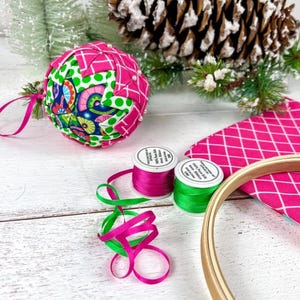 May include: A quilted fabric ornament featuring a geometric pattern of pink, green, and blue floral fabric, accompanied by two spools of 4mm pure silk ribbon in pink and green, a piece of pink diamond-patterned fabric, and a wooden embroidery hoop.