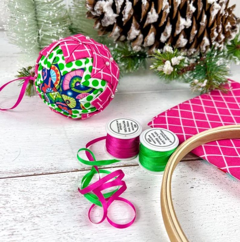 May include: A quilted fabric ornament featuring a geometric pattern of pink, green, and blue floral fabric, accompanied by two spools of 4mm pure silk ribbon in pink and green, a piece of pink diamond-patterned fabric, and a wooden embroidery hoop.