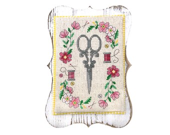 May include: A framed cross-stitch featuring a pair of scissors, spools of thread, and pink flowers. The design is surrounded by a floral wreath and set against a beige background with a yellow border. The frame is white with a distressed finish.