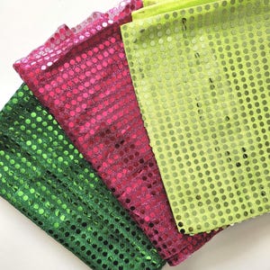 May include: Close-up of three sequined fabric swatches in green, fuchsia, and lime green. The round sequins are densely packed, creating a shimmering effect. The fabric is folded and layered, showing the texture and colour variations.