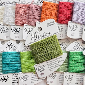 Pode incluir: Uma coleção de fios de bordar Glisten Sparkling Wool Braid em cartões brancos. Cada cartão contém 11 metros de uma mistura de 65% lã merino e 35% poliéster metalizado em várias cores, incluindo branco, rosa, laranja, vermelho, roxo e verde.