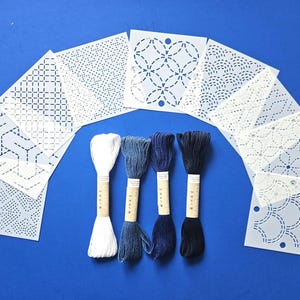 May include: A collection of nine square white stencils featuring various geometric patterns arranged in a fan shape, accompanied by four skeins of embroidery thread in shades of white, light blue, medium blue, and dark navy blue.