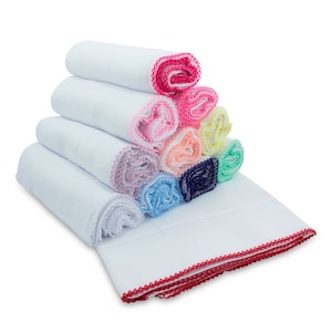 Op de afbeelding: Een stapel witte keukendoeken met kleurrijke gehaakte randen. De handdoeken zijn gevouwen en gestapeld in een piramidevorm. De bovenste handdoek heeft een rode gehaakte rand, de volgende handdoek heeft een roze gehaakte rand, enzovoort. De handdoeken zijn allemaal wit en hebben een zachte, absorberende textuur.