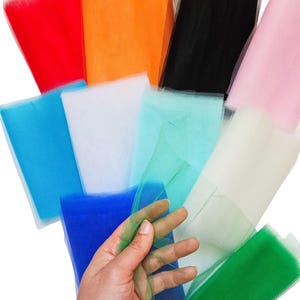 Könnte beinhalten: Eine Sammlung farbenfroher Tüllstoffrollen in verschiedenen Farbtönen, darunter Rot, Orange, Schwarz, Rosa, Blau, Weiß und Grün. Eine Hand hält ein Stück grünen Tüll, das die Textur und Farbe des Stoffes zeigt. Ideal für Bastel- und Dekorationsprojekte.