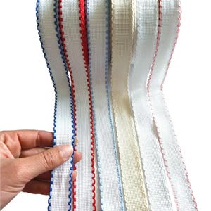 Puede incluir: Cinta blanca tejida con bordes festoneados en varios colores, incluyendo rojo, azul y crema. La cinta se sostiene en una mano, mostrando las diferentes opciones de color. Ideal para proyectos de manualidades y costura.