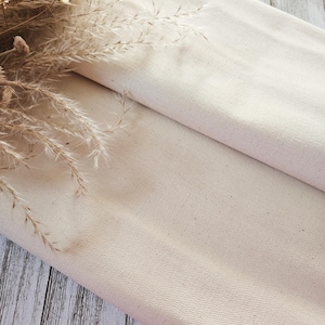 May include: Folded, off-white canvas fabric with a textured surface. Dried, beige-coloured ornamental grasses and flowers are arranged in the upper left corner. The fabric is draped over a light-coloured wooden surface.