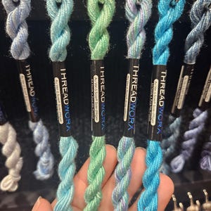 May include: A selection of colourful embroidery thread spools in shades of blue, green, and purple. Each spool is labelled with the brand "THREAD WORX" and includes text in French. The spools are displayed on a metal hook.