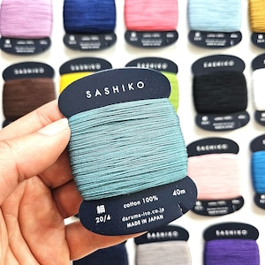 May include: A teal Sashiko embroidery thread wound on a black cardboard bobbin, labelled 100% cotton, 40m, 20/4, and made in Japan. The bobbin features the text SASHIKO and daruma-ito.co.jp, surrounded by other bobbins in various colours.