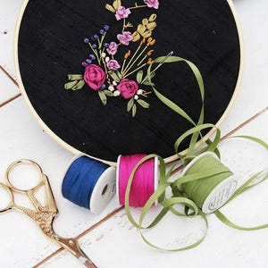 May include: A round wooden embroidery hoop containing black fabric with floral ribbon embroidery in shades of pink, yellow, and blue. Three spools of ribbon in blue, magenta, and olive green sit nearby with a pair of gold-toned metal scissors.