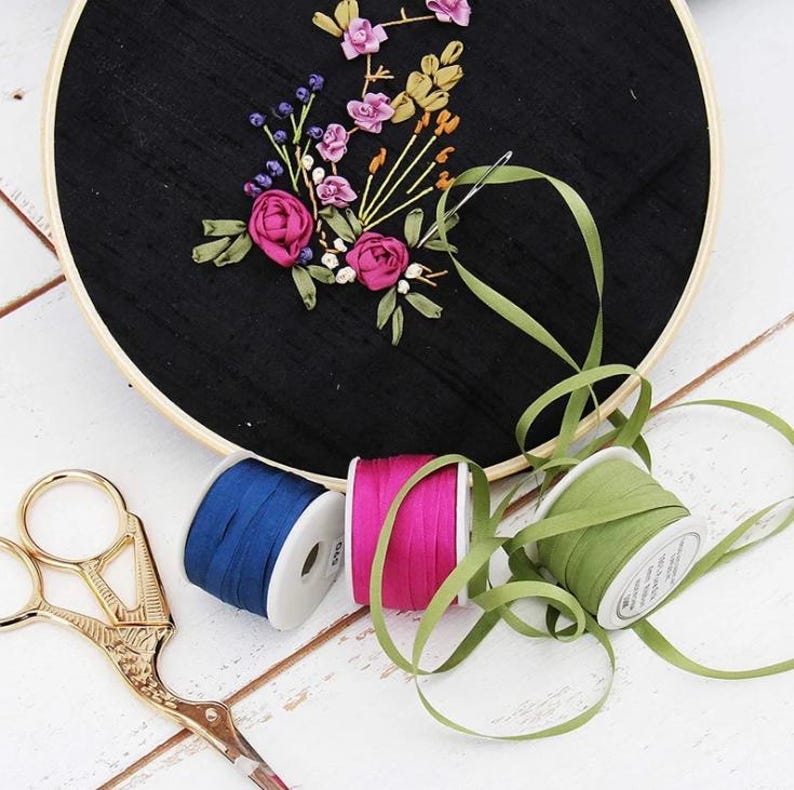 May include: A round wooden embroidery hoop containing black fabric with floral ribbon embroidery in shades of pink, yellow, and blue. Three spools of ribbon in blue, magenta, and olive green sit nearby with a pair of gold-toned metal scissors.
