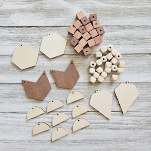 Puede incluir: Una colección de suministros de artesanía de madera sin terminar, que incluye formas geométricas, cubos y cuentas. Las formas incluyen hexágonos, diamantes y semicírculos, todos con pequeños agujeros para ensartar. Las piezas de madera están dispuestas sobre una superficie clara y texturizada.
