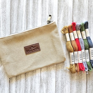 Puede incluir: Una bolsa de lona beige con cremallera y un parche de cuero marrón que dice "DMC ECO VITA". Junto a ella hay varios carretes de hilo de bordar de colores en tonos amarillos, rojos, azules y verdes. Los artículos están dispuestos sobre una superficie de madera clara.