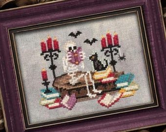 May include: Cross-stitch artwork in a purple frame. The image features a skeleton, black cat, and candles on a stack of books, with bats flying above. The color palette includes red, black, white, and brown, creating a Halloween theme.