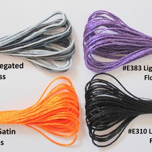 May include: Four spools of embroidery floss in different colors and textures. The top left spool is gray and white variegated floss, labeled #53 Variegated Floss. The top right spool is purple with a shimmery finish, labeled #E383 Light Effects Floss. The bottom left spool is orange satin floss, labeled #S741 Satin Floss. The bottom right spool is black with a matte finish, labeled #E310 Light Effects Floss.