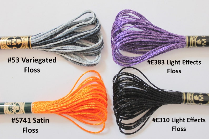 May include: Four spools of embroidery floss in different colors and textures. The top left spool is gray and white variegated floss, labeled #53 Variegated Floss. The top right spool is purple with a shimmery finish, labeled #E383 Light Effects Floss. The bottom left spool is orange satin floss, labeled #S741 Satin Floss. The bottom right spool is black with a matte finish, labeled #E310 Light Effects Floss.