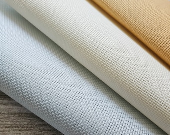 May include: Three rolls of Aida cloth in white, light blue, and beige. The cloth is a woven fabric with a grid pattern, commonly used for cross-stitch embroidery.