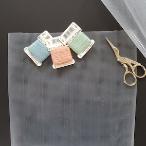 May include: Three spools of embroidery floss in shades of light blue, peach, and green. The spools are labeled with the brand "Anchor" and the color numbers 3901, 3916, and 7458. The spools are on a gray plastic canvas sheet.