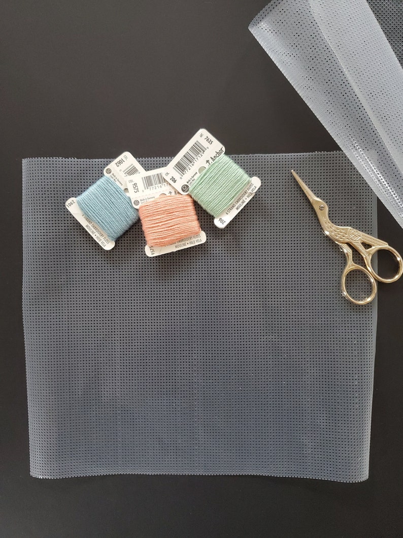 May include: Three spools of embroidery floss in shades of light blue, peach, and green. The spools are labeled with the brand "Anchor" and the color numbers 3901, 3916, and 7458. The spools are on a gray plastic canvas sheet.