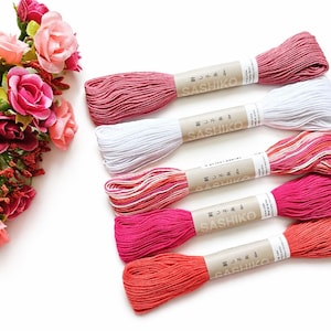 May include: Six spools of Sashiko thread in shades of pink, white, and red are arranged on a white surface. The spools are labelled with Japanese text. A bouquet of pink and red roses is in the upper left corner.