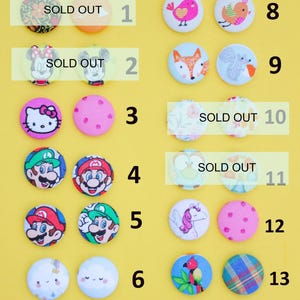 a collection of 14 colorful buttons, each with a unique design, arranged in two rows against a yellow background. The buttons are labeled with numbers 1 through 14, indicating that they are available for purchase.