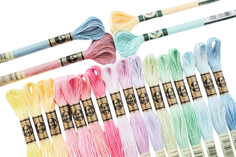 May include: A collection of colorful embroidery floss skeins in various pastel shades, including yellow, pink, blue, and green. Each skein is labeled with "M C 25" on a black and gold label, indicating the thread type and weight. Ideal for embroidery and cross-stitch projects.