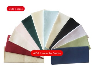 Può includere: Un ventaglio di campioni di tessuto AIDA 11 count in vari colori, tra cui rosso, crema, verde e blu. Il testo "Made in Japan" e "AIDA 11 count by Cosmo" sono visibili.