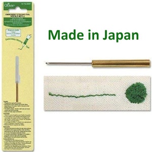 May include: A close-up of a gold embroidery stitching tool with a needle refill. The tool is made in Japan and is shown on a white fabric with green stitching. The packaging for the tool is shown on the left side of the image.