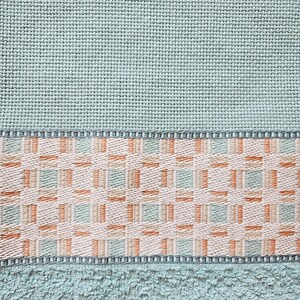 May include: Close-up of a light teal towel with a woven geometric pattern in the center. The towel has a textured surface and a border with a repeating design. The color palette includes teal, beige, and orange.