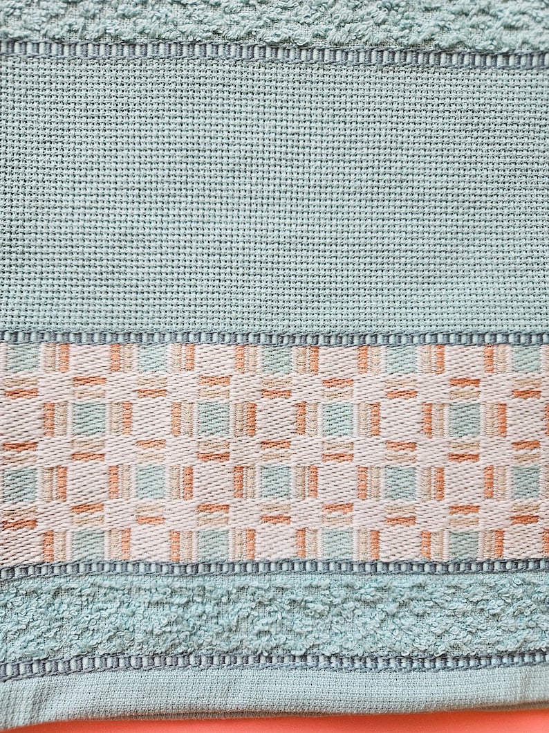 May include: Close-up of a light teal towel with a woven geometric pattern in the center. The towel has a textured surface and a border with a repeating design. The color palette includes teal, beige, and orange.