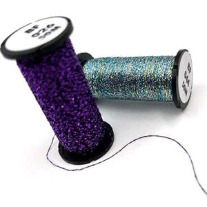 Pode incluir: Duas bobinas de fio decorativo. Uma é roxa vibrante com textura brilhante, a outra é turquesa cintilante. Cada bobina tem base preta e etiqueta branca com texto preto. O fio tem aproximadamente 50 metros de comprimento.
