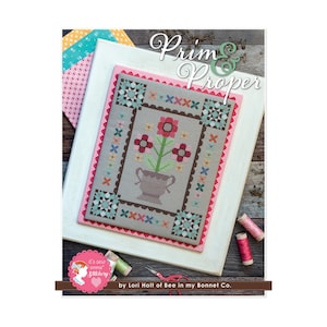 Peut inclure: Un modèle de point de croix intitulé "Prim & Proper" représentant un motif floral avec un vase et des fleurs dans des tons de rouge, rose et vert. Le motif est encadré dans un cadre blanc avec une bordure en vichy rose et blanc.