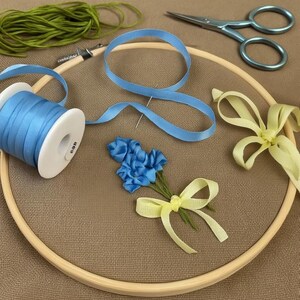 May include: A wooden embroidery hoop containing a brown fabric base with a blue ribbon flower embroidery, a spool of blue ribbon, a loose blue ribbon, a pale yellow ribbon bow, green embroidery thread, and blue metal scissors.