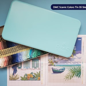 May include: A light blue tin with the text "DMC Scenic Colors Tin 35 Skeins" on the lid. The tin is open and contains 35 skeins of embroidery floss in various colours. A cross-stitch pattern is visible in the background.