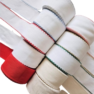 May include: A selection of white fabric ribbons with decorative edges and coloured trim. The ribbons are rolled and stacked, featuring red, green, blue, and gold trim. The ribbons have a textured weave.