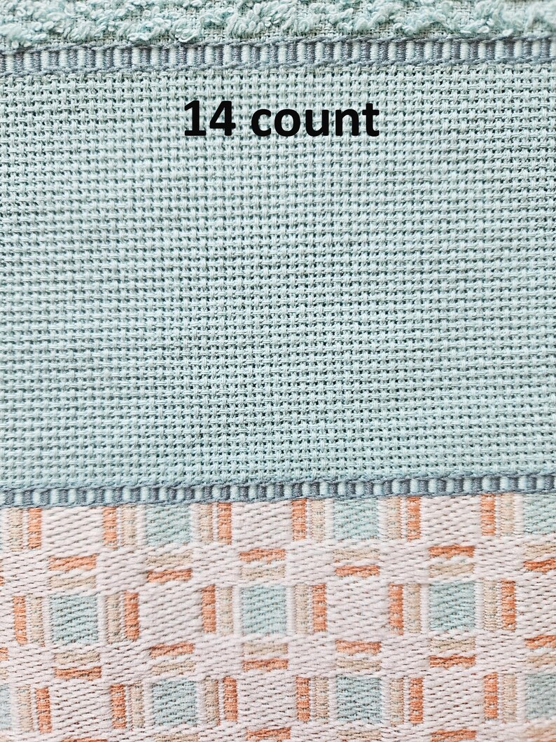 May include: Close-up of a section of cross-stitch fabric. The top portion is a light teal color with the text "14 count" in black. The bottom section features a woven pattern in white, peach, and teal.