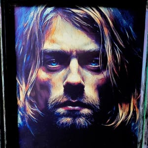 Puede incluir: Retrato enmarcado de una persona con pelo largo y claro y barba. El retrato tiene ojos azules y una expresión seria. El marco es negro desgastado con detalles turquesa, lo que le da un aspecto vintage.