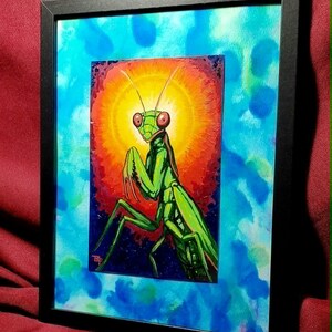 Puede incluir: Una obra de arte enmarcada que presenta una mantis religiosa de color verde vibrante centrada sobre un fondo circular de color naranja y amarillo. El borde circundante muestra un estilo de acuarela azul y verde azulado, en un marco negro.