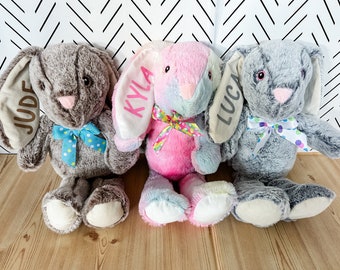 May include: Three plush bunny toys with personalized names on their ears. The bunnies are brown, pink, and gray with colorful bows around their necks. The names on the bunnies are Jude, Kyla, and Luca.