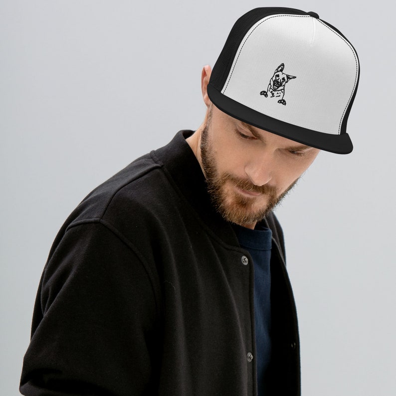 May include: A black and white trucker hat with a black mesh back. The front of the hat features an embroidered image of a dog peeking out from behind the brim.