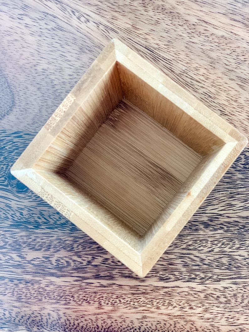May include: A square wooden box with a natural wood finish. The box is empty and has a smooth, polished surface.