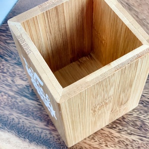 May include: A small, square, wooden container made of bamboo. The container is empty and has a light brown colour.