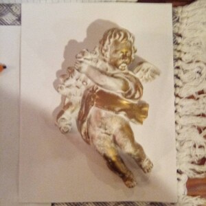 May include: A gold-colored cherub figurine with outstretched arms, holding a bouquet. The cherub has detailed wings and is set against a white background. The figurine has a vintage, antique appearance.