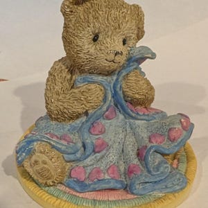 May include: A small, tan-colored teddy bear figurine holding a blue blanket decorated with pink hearts. The bear sits on a round, pastel-colored base. The figurine is made of a textured material.