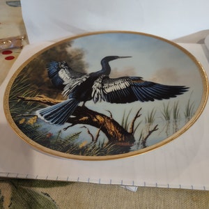 May include: A decorative ceramic plate with a gold rim features a painting of a blue and black bird with white markings perched on a branch. The bird has its wings spread out and is surrounded by green foliage.