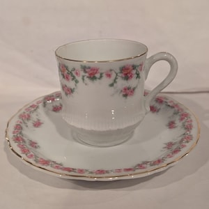 May include: A white teacup and saucer set with pink rose floral designs and gold trim. The teacup has a handle and a ribbed lower section. The saucer has a scalloped edge. A classic tea set.
