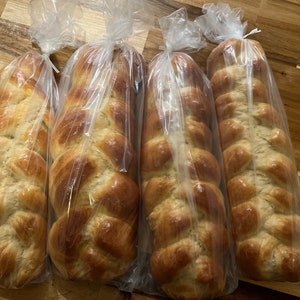 May include: Four braided loaves of golden-brown challah bread, each wrapped in a clear plastic bag. The bread has a glossy finish and is displayed on a wooden surface.
