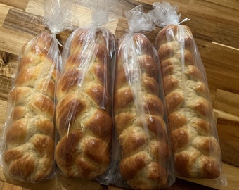 May include: Four braided loaves of golden-brown challah bread, each wrapped in a clear plastic bag. The bread has a glossy finish and is displayed on a wooden surface.