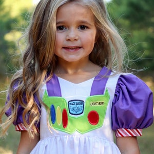 May include: A white dress with purple accents and a green chest piece, resembling a Space Ranger uniform. The dress features the text "LIGHTYEAR" and "SPACE RANGER". The sleeves and cuffs have red and white stripes. The dress is for children.